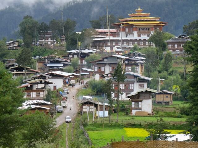 Gangtey Goemba monastery visible in the distance, overlooking a valley with scattered houses, green fields, and mountains under a clear sky.