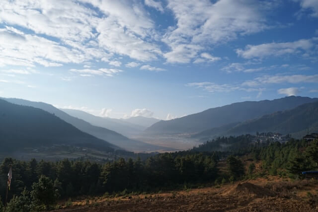 Phobjikha Valley under a clear blue sky, surrounded by rolling green hills, dense trees, and distant mountain ranges creating a serene natural landscape.