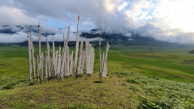 Phobjikha Valley featuring wide green open fields with white prayer flags fluttering in the wind, set against surrounding hills and distant mountains under a clear sky.